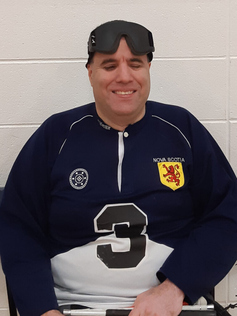 A man smiles, wearing a blue & white jersey with the number 3 and Nova Scotia heraldic lion. Goalball eyeshades sit on top of his head. 